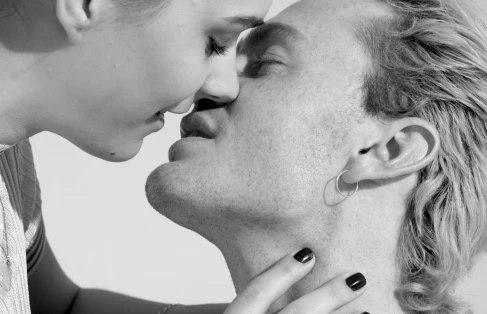 Close-up black and white portrait of two models kissing, featuring dark nail polish, silver hoop earring, and clean background