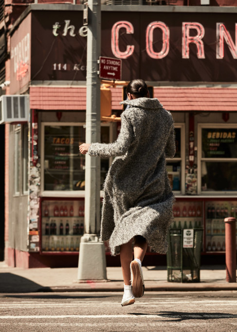 Woman with long coat crossing street near The Corner restaurant in New York City on a sunny day, back view. Fashion editorial.