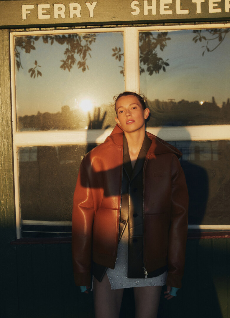 A woman in a leather jacket stands in front of a ferry shelter, bathed in warm, golden light from the setting sun, fashion shot.