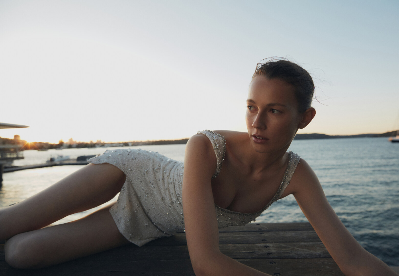 Woman in beaded dress lying on wooden dock with water and cityscape in background during golden hour for fashion or beauty campaign.