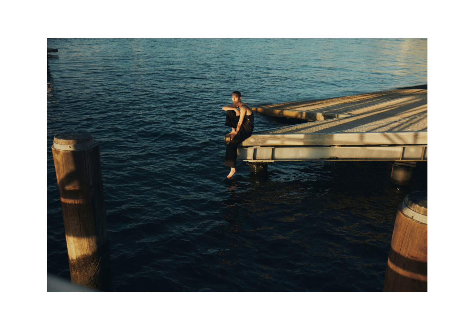 Adult female in black outfit sitting on a dock with legs dangling above the water, enjoying the scenery with a contemplative expression on her face.