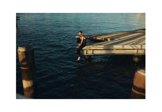 Adult female in black outfit sitting on a dock with legs dangling above the water, enjoying the scenery with a contemplative expression on her face.