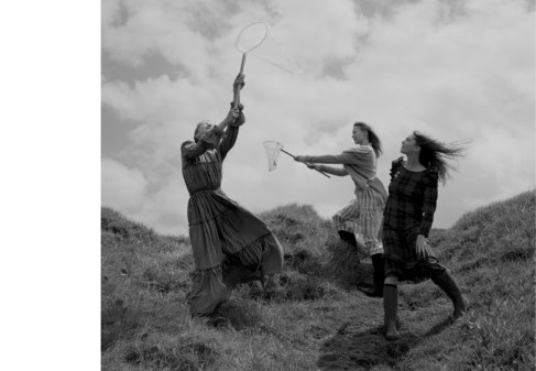 Three women stand on a grassy hill, holding nets, wearing long dresses, in a black and white outdoor shot under a cloudy sky.