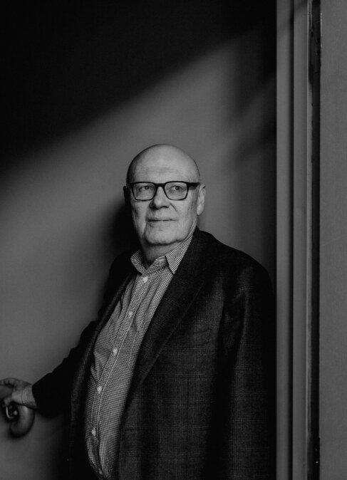 Portrait of an older man in glasses wearing a blazer and patterned shirt posing indoors near a doorway
