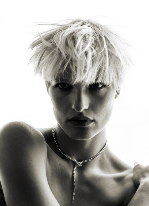 Portrait of a woman with platinum blonde hair wearing a snake necklace against a bright background in a monochrome photograph.