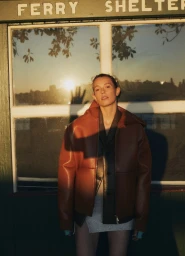 Model Mali Koopman in a rust leather jacket standing against a ferry shelter window during sunset for Human Story by Side-note.
