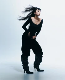Model with long dark hair blowing back wearing a black bodysuit and slouchy boots against a white backdrop.