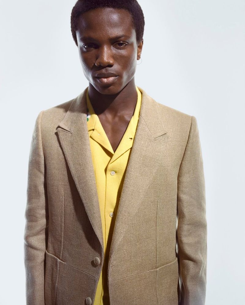 Close-up of an adult Black male in a tan blazer over a yellow shirt, against a white backdrop. Fashion shot, professional portrait.