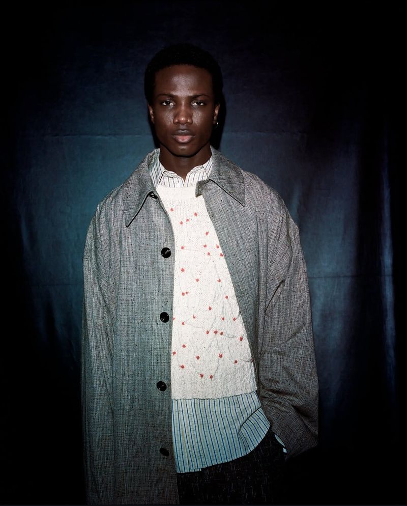 Studio shot of an adult Black man wearing a grey overcoat and patterned top, standing against a dark blue backdrop in portrait orientation.