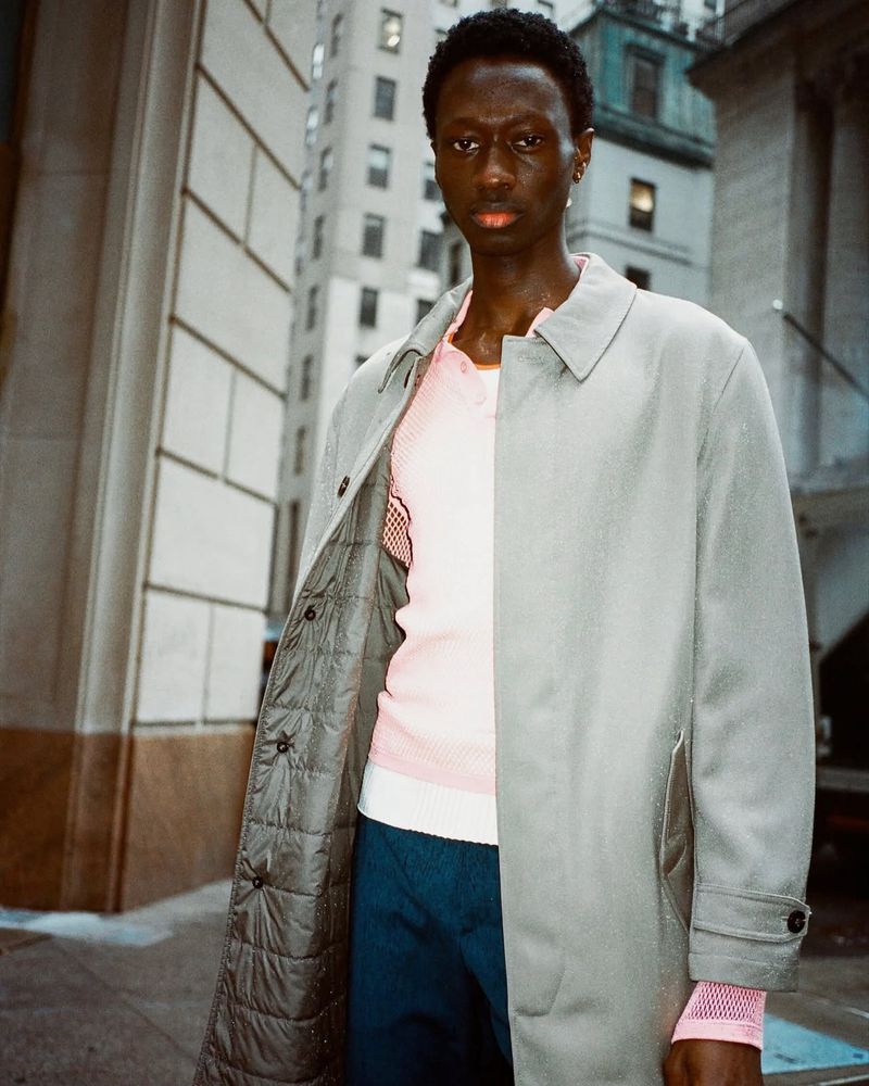 Man in gray coat and pink shirt posing in city street. Tall buildings and architectural detail visible in background. Fashion editorial.