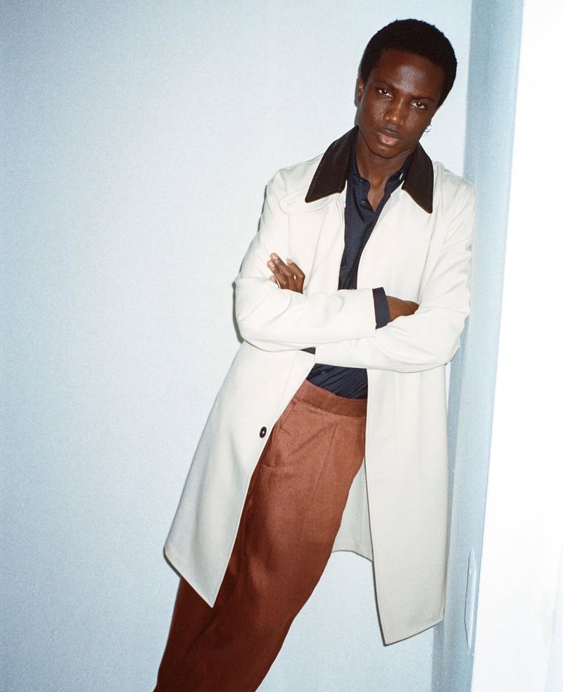 Young Black man posing with arms crossed in a studio setting, wearing a cream overcoat, brown slacks, and a navy shirt with dark brown collar.