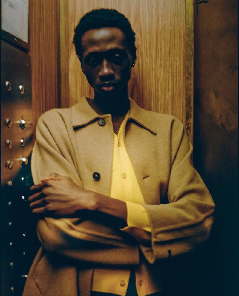 Portrait of a black male model in a tan overcoat and bright yellow shirt in an elevator with his arms crossed, looking at the camera.