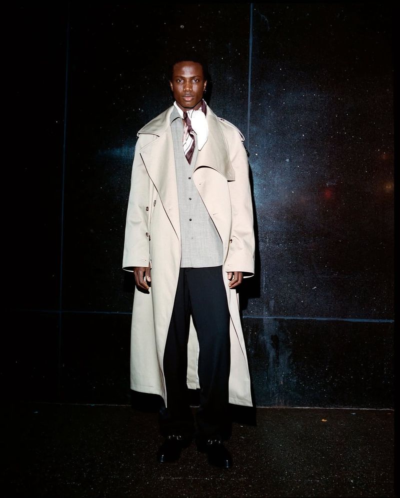 A man wearing a tan trench coat and patterned scarf posing against a dark wall for an editorial shoot.