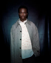 Model in grey coat and patterned sweater against a dark backdrop for T The New York Times Style Magazine