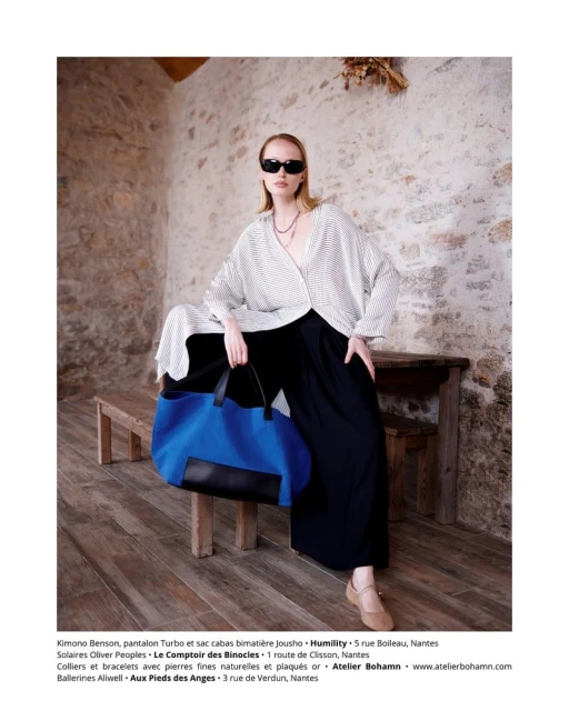 Fashion shot featuring an adult woman in a loose white top and black pants, accessorized with sunglasses and a necklace, posing indoors.