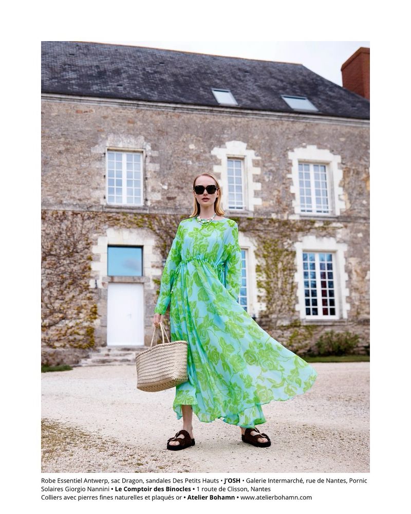Woman in a flowing floral dress and sandals stands in front of a stone building with straw bag, sunglasses, and jewelry.