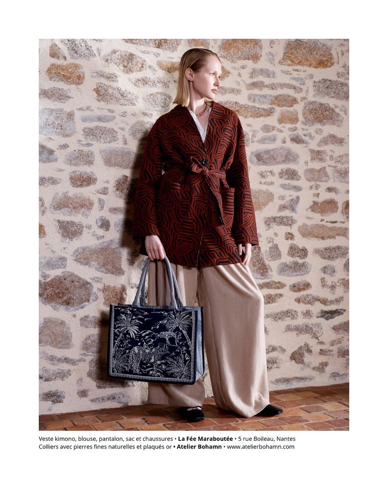 A woman models a kimono-style jacket with a patterned bag and wide-legged trousers in front of a textured stone wall indoors.
