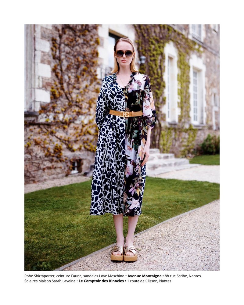 Fashion shot of a woman wearing a floral dress, sunglasses, and platform sandals, standing on a stone path in front of a stone building.