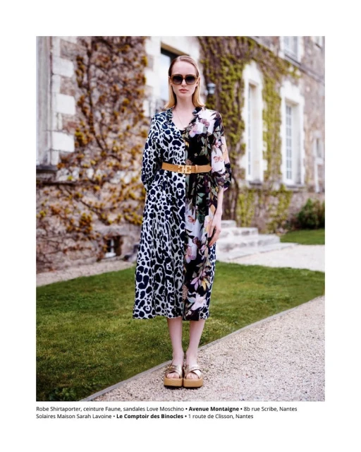 Fashion shot of a woman wearing a floral dress, sunglasses, and platform sandals, standing on a stone path in front of a stone building.