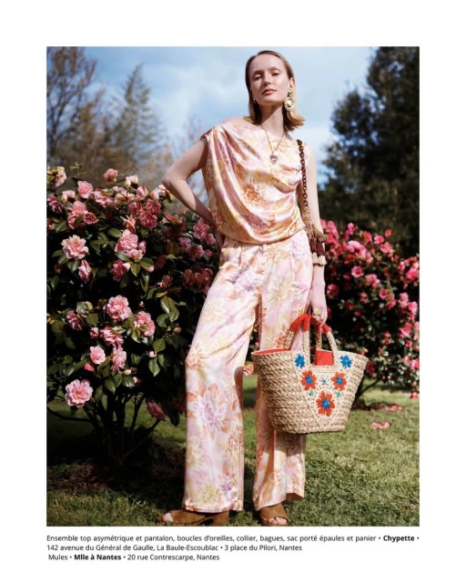 Woman in floral blouse and pants poses outdoors next to flowers with straw tote bag featuring floral embroidery and embellishments.