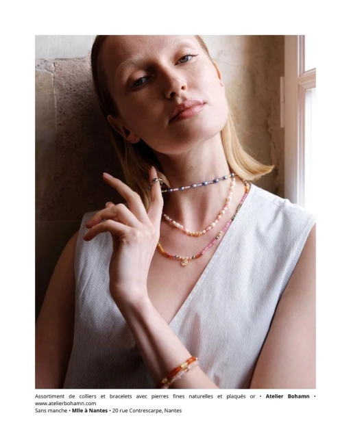 Woman with blonde hair wearing layered pearl necklaces and bracelet, posing indoors with natural light near a window. Jewelry fashion.