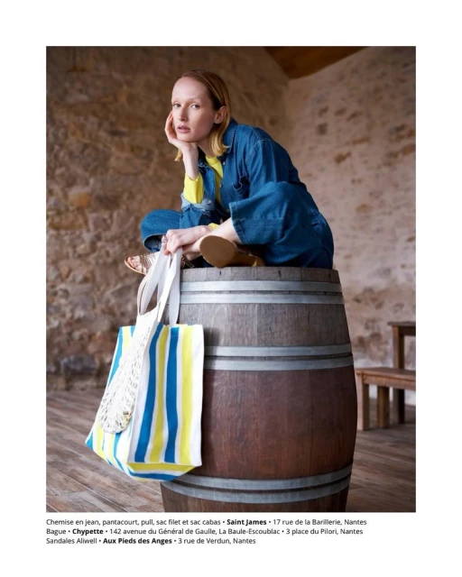 Adult female in blue denim jumpsuit seated on a wooden barrel in a rustic indoor setting, holding a striped tote bag, styled hair, neutral expression.