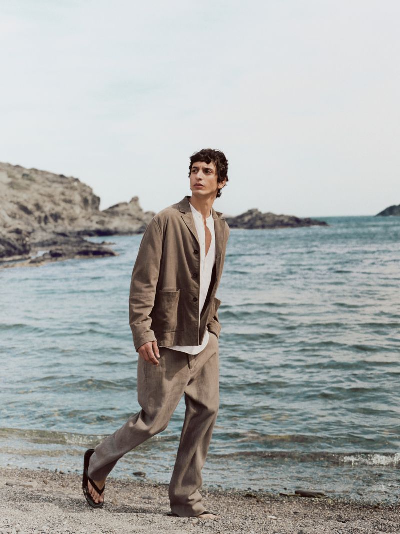 A male model wearing a taupe jacket and trousers walking on a sandy beach by the ocean for Massimo Dutti Tideborn.