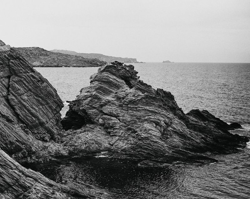 Black and white scenic shot of rugged, layered rock formations meeting the ocean under an overcast sky, creating a stark coastal landscape.