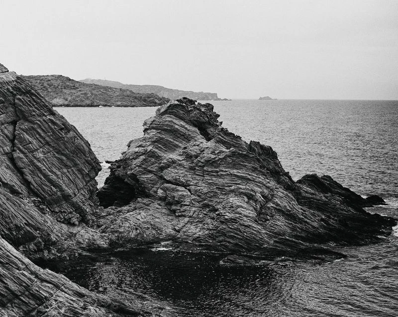 Black and white scenic shot of rugged, layered rock formations meeting the ocean under an overcast sky, creating a stark coastal landscape.