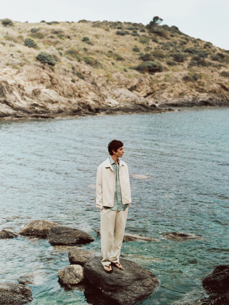 A man stands on rocks in the ocean wearing a light jacket and pants, with a grassy hill in the background on a cloudy day.