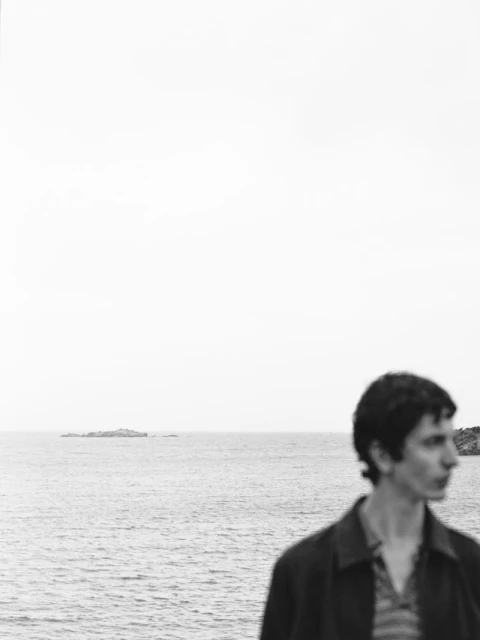 Portrait of a young man standing outdoors near the ocean with a dark jacket and striped shirt, captured in black and white.