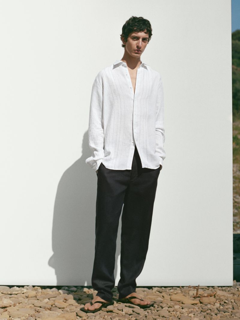 A young male model stands against a white wall outdoors, wearing a white linen shirt and dark trousers in a casual, fashion editorial style.