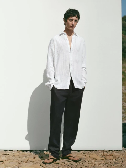 A young male model stands against a white wall outdoors, wearing a white linen shirt and dark trousers in a casual, fashion editorial style.