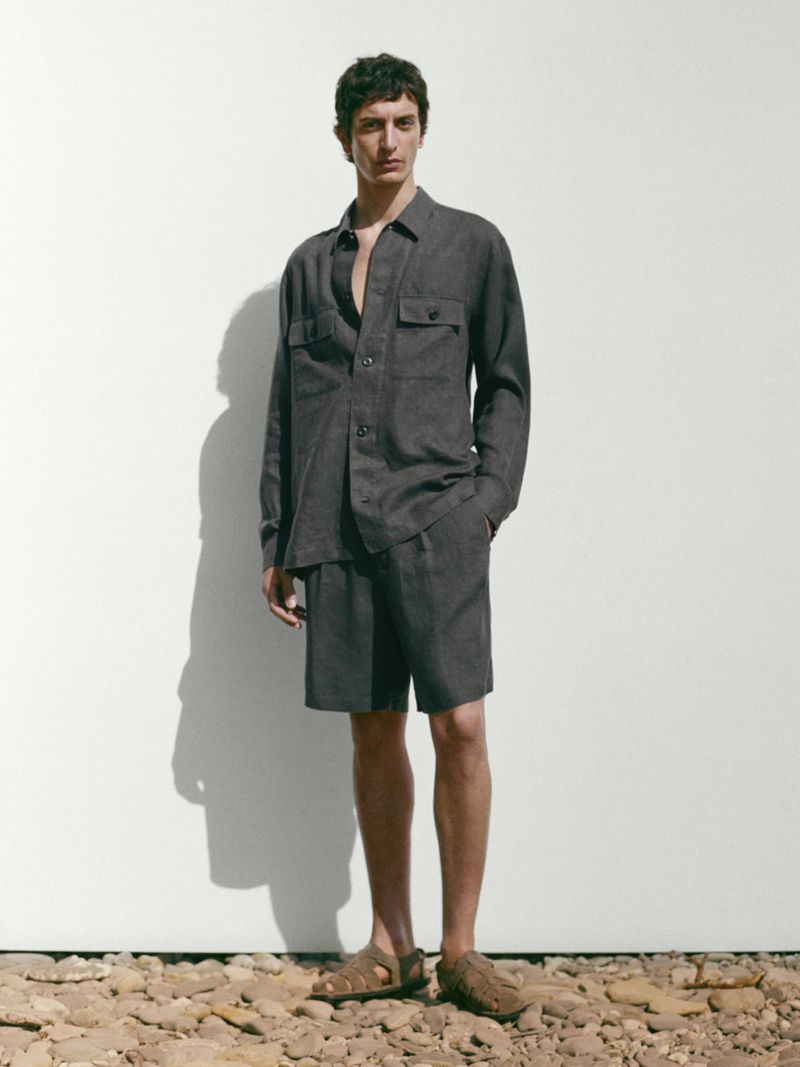 An adult male models a brown button-down shirt and shorts outfit with leather sandals against a white wall in a studio setting.