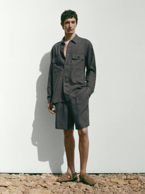An adult male models a brown button-down shirt and shorts outfit with leather sandals against a white wall in a studio setting.