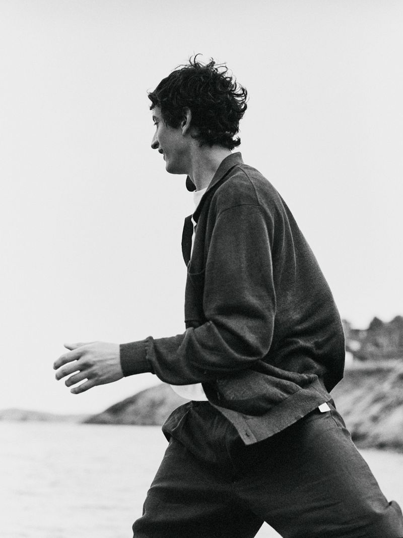 Monochrome medium shot of a man outdoors, side view, wearing a cardigan, dark pants, and white undershirt. Water and land in the background.