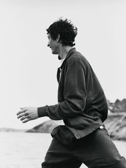 Monochrome medium shot of a man outdoors, side view, wearing a cardigan, dark pants, and white undershirt. Water and land in the background.