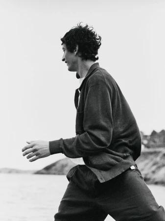 Monochrome medium shot of a man outdoors, side view, wearing a cardigan, dark pants, and white undershirt. Water and land in the background.