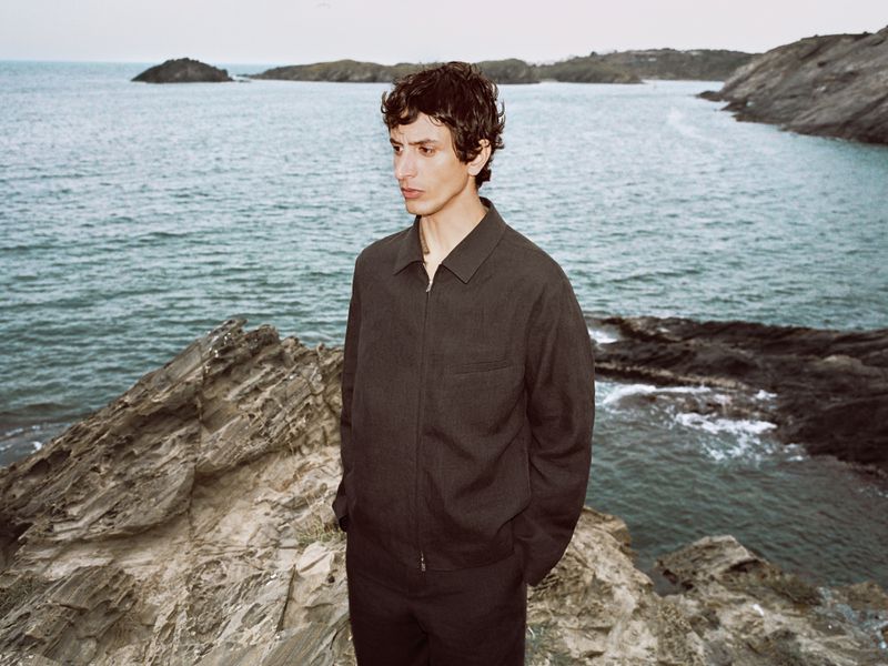 Model wearing a dark jacket standing on rocky coastline overlooking the ocean for Massimo Dutti