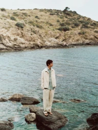 A man standing on a rock by the ocean wearing a light-coloured jacket and trousers with a rocky hillside behind him.