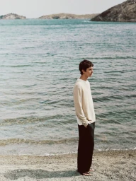 Man wearing a cream knit sweater and black trousers standing by the ocean shore on a sandy beach