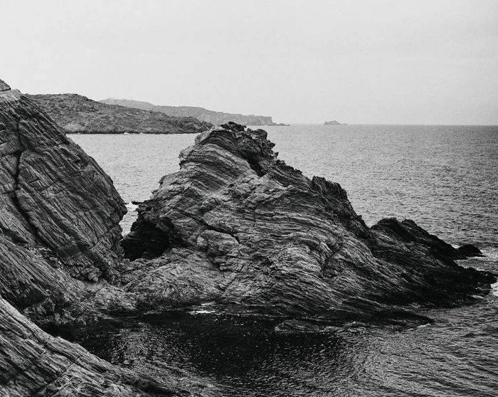 Monochrome landscape photograph of jagged rocky cliffs meeting the dark open ocean under an overcast sky.