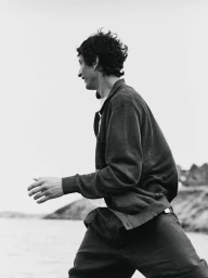 Black and white portrait of a male model wearing a dark zipped jacket outdoors near a coast by Ami Lusinchi