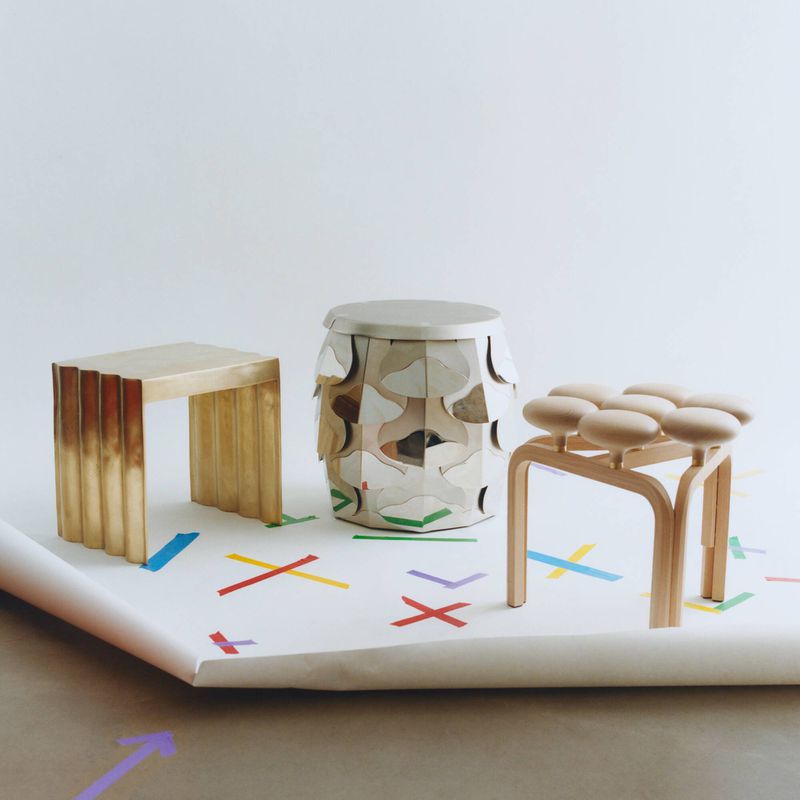 Three distinctive design objects including a gold metallic side table, a mirrored cylindrical stool, and a wooden multi-lobed stool.