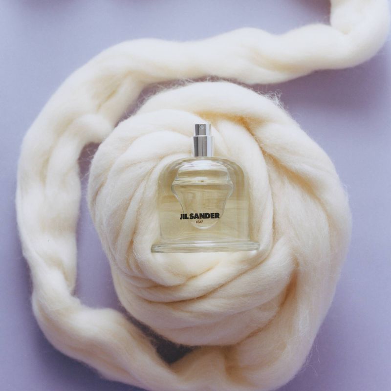 A bottle of Jil Sander perfume sits nestled in a bed of white wool against a light purple background in a studio shot.