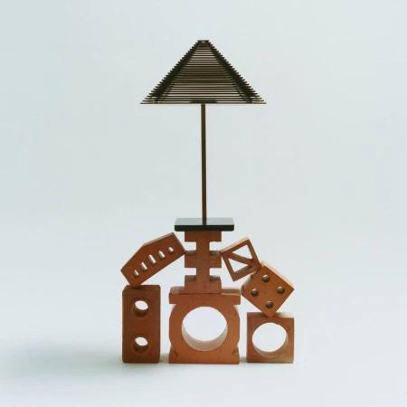 A sculptural arrangement of terracotta blocks and perforated bricks topped with a slender wire pyramid structure on a white seamless background.