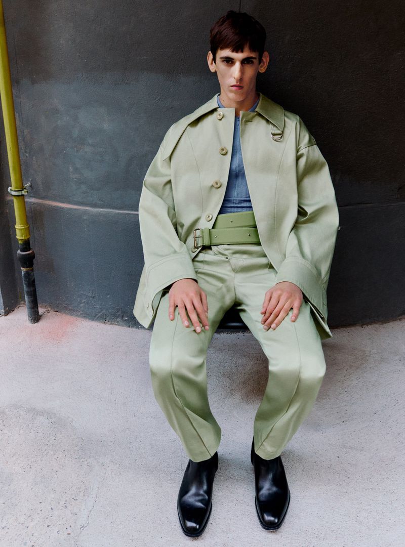 A fashion shot of a young adult male model wearing a light green matching coat and pants, black boots, and a blue shirt against a gray wall.