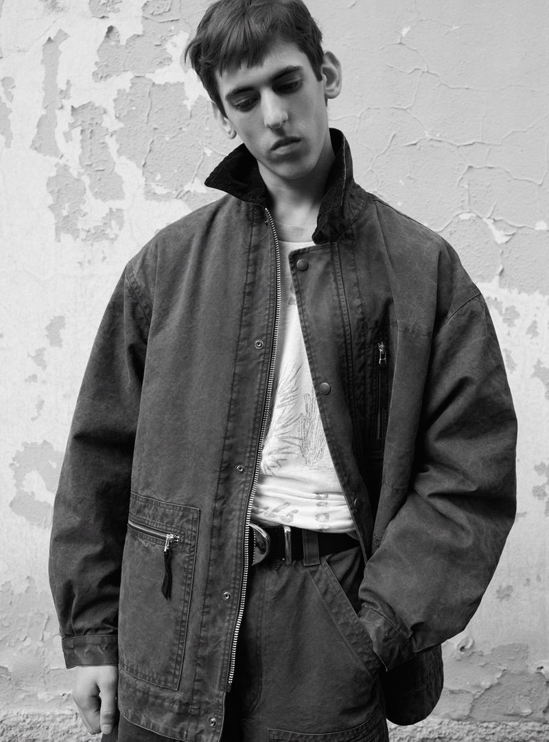 Monochrome medium shot of a young adult male wearing a jacket, t-shirt, and pants in front of a weathered wall, looking downward.