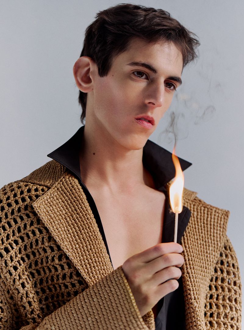 Young man in a tan, woven jacket holds a lit match in a studio setting against a neutral background, showcasing fashion-forward style.