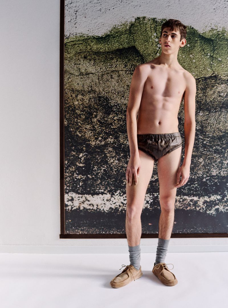 A shirtless young man in dark briefs, grey socks, and tan shoes stands indoors in front of a mottled earth-tone background poster.
